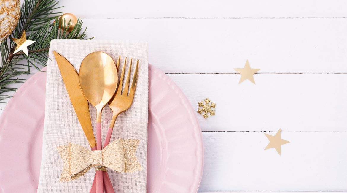 A pink and gols ilverware set sits on top of a pink dinner plate. The background is decorated for Christmas.